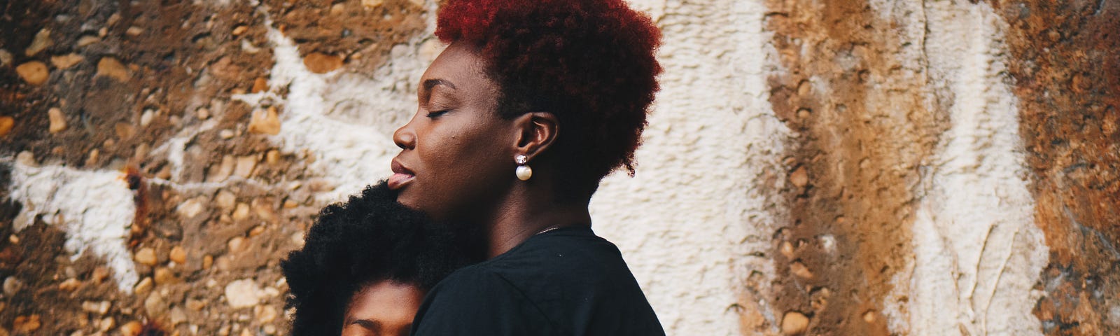 Black mother and daughter hug in front of a wall.