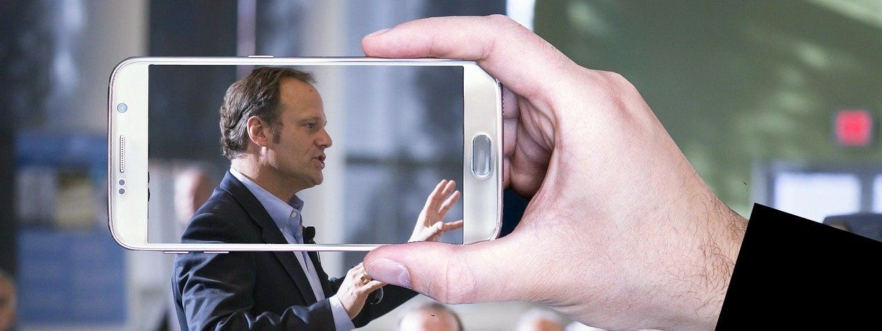 Man speaking in public while someone snaps his picture with a smartphone