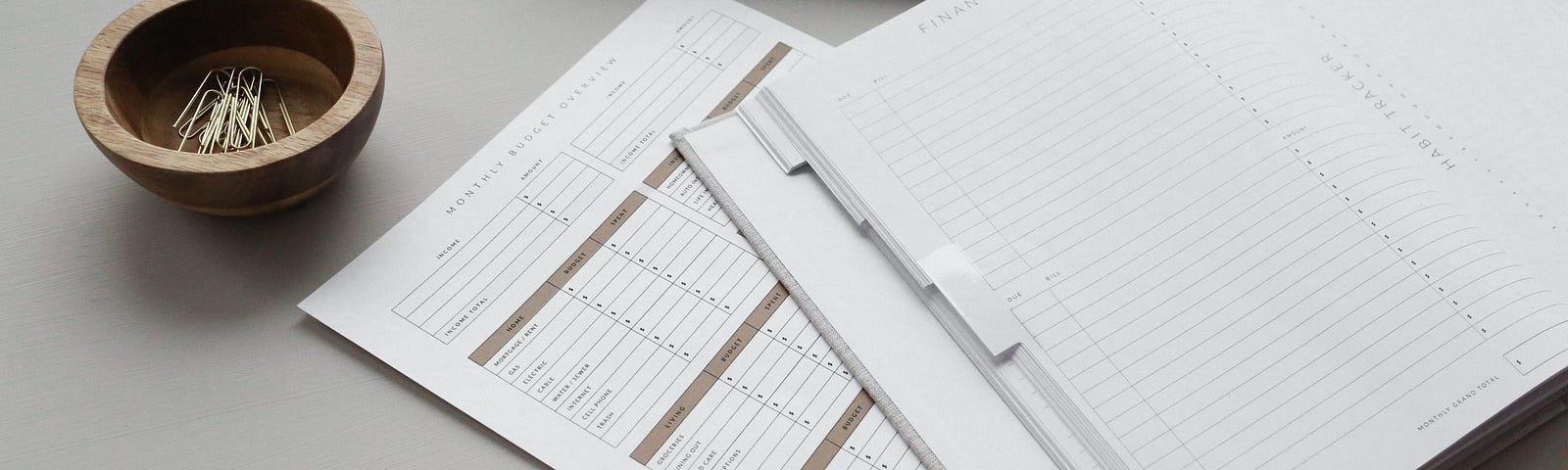 Paper budget worksheet, finance tracking notebook, pencil, and tray of paperclips on a gray background.