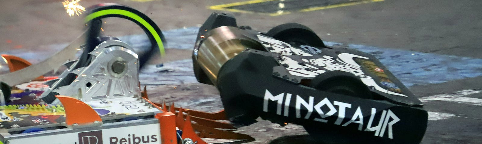 A weapon-on-weapon hit between End Game and Minotaur. End Game is a white and orange box-bot with a vertical spinner. Orange forks jut fro the front. Minotaur is a black box with white lettering and a while minotaur painted on the top. A wide vertical drum spinner sits at the front of the bot.