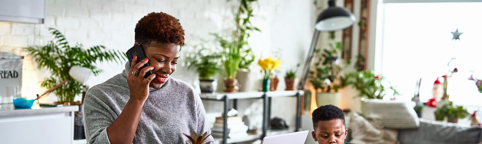 An adult woman on the phone looking at her laptop on the dining table while her son uses a digital tablet.