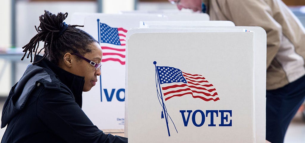 A photo of a black woman voting at a poll. There is a divider on the desk that reads “VOTE” with a U.S. flag.