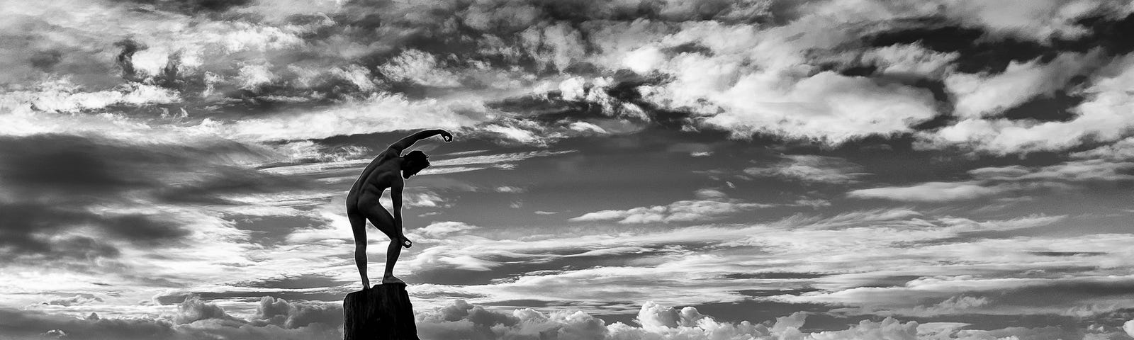 A nude man stands on a tree stump on the beach, the Pacific Ocean behind him with an amazing array of spotted and dramatic clouds filling the sky while he balances in a yoga’esque pose.