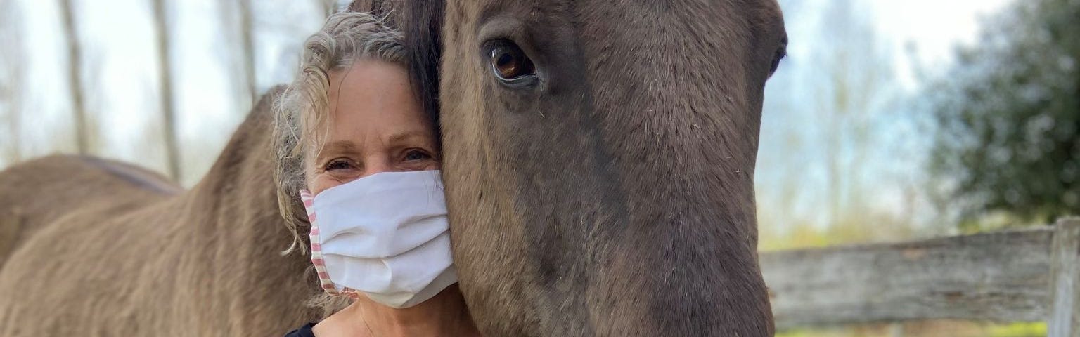 Keryn Denroche, a woman with short gray hair and a gray mask, poses with a soft looking brown horse on her farm sanctuary