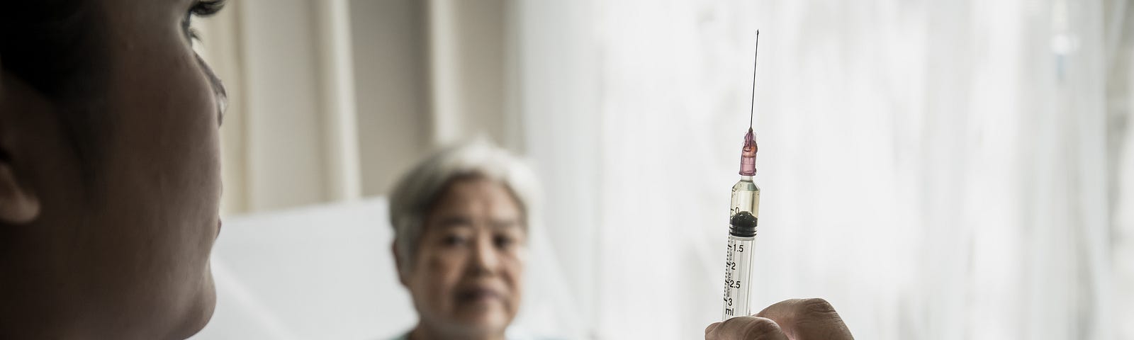 A photo of a nurse holding up a syringe with an elderly woman sitting in a hospital bed in the background.