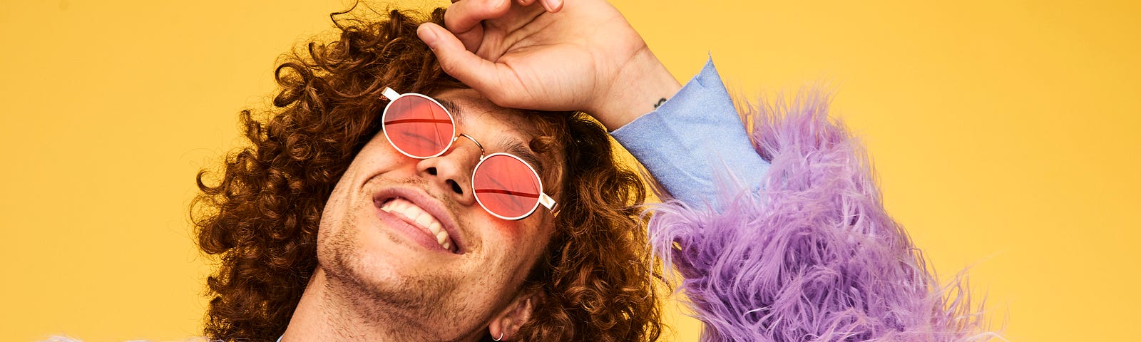 A photo of a joyful man with curly brown hair wearing a lavender furry jacket in front of a yellow background.