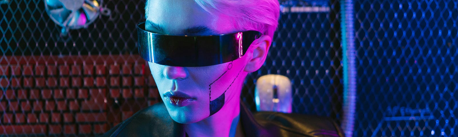 A woman with short, blond hair, poses in  black futuristic eyewear. She’s wearing a black leather jacket with symmetrically lined facial makeup on her cheek. There is a red keyboard behind a mesh fence in the background, and the image has a slight pink glow.