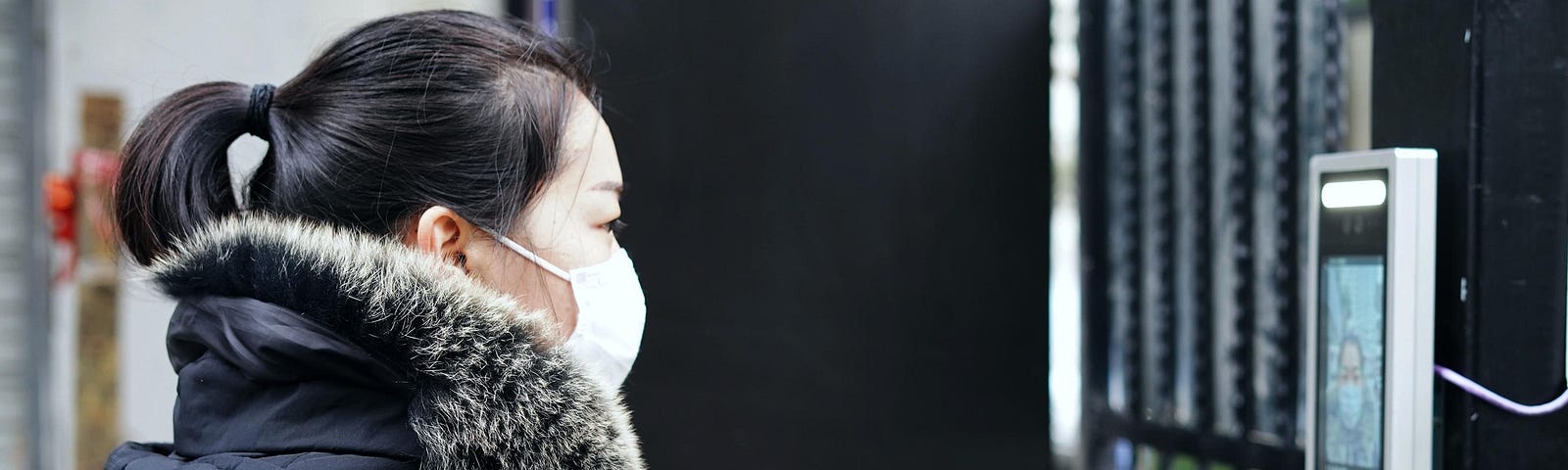 A woman with a face mask uses a facial recognition system to enter a residence.