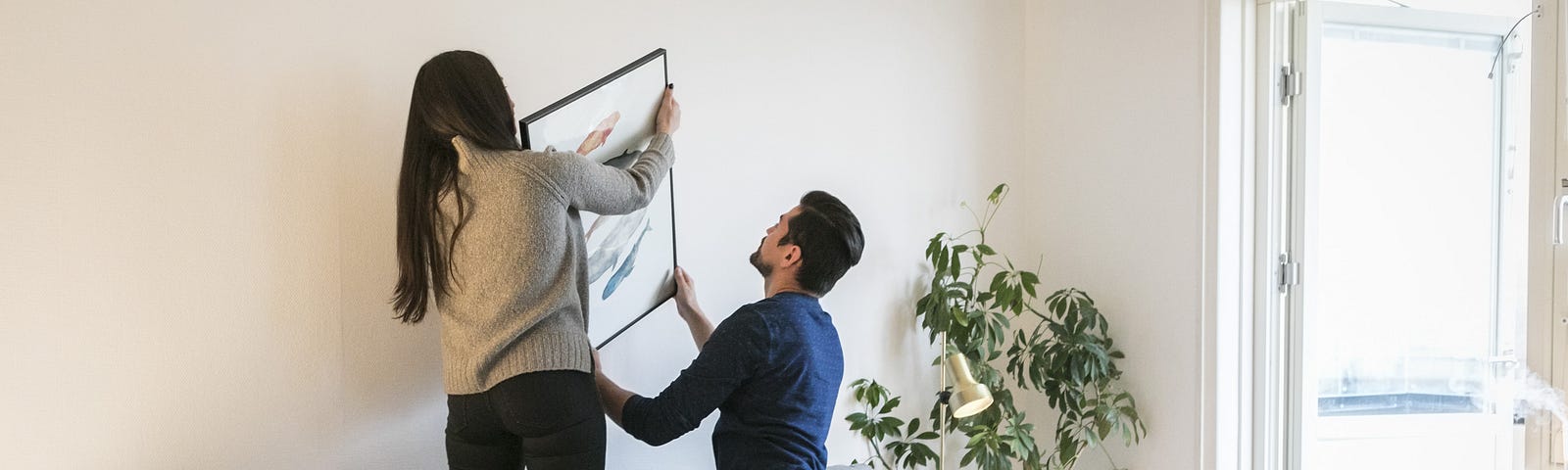 A couple hanging a framed picture on their living room wall.