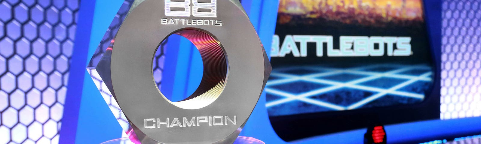 A picture on the Giant Nut, the trophy for BattleBots. It’s a literal steel hexagonal nut about 8 inches across. One one side the BattleBots logo is etched above the hole, and Champion is etched below the hole. It sits on a glass disk, which sits on a five-finger robotic pedestal. The finger manually rotate the glass disk.