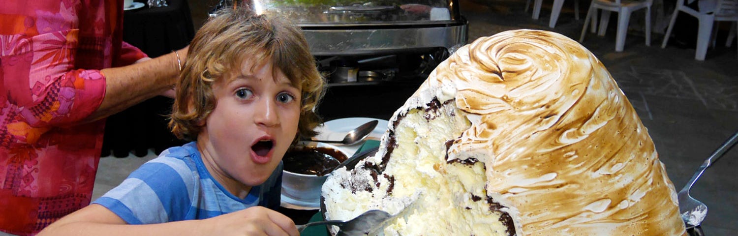 A young fair-haired boy in a two-toned blue striped t-shirt has his mouth wide open in a look of surprise as he scoops into an enormous Bombe Alaska dessert.