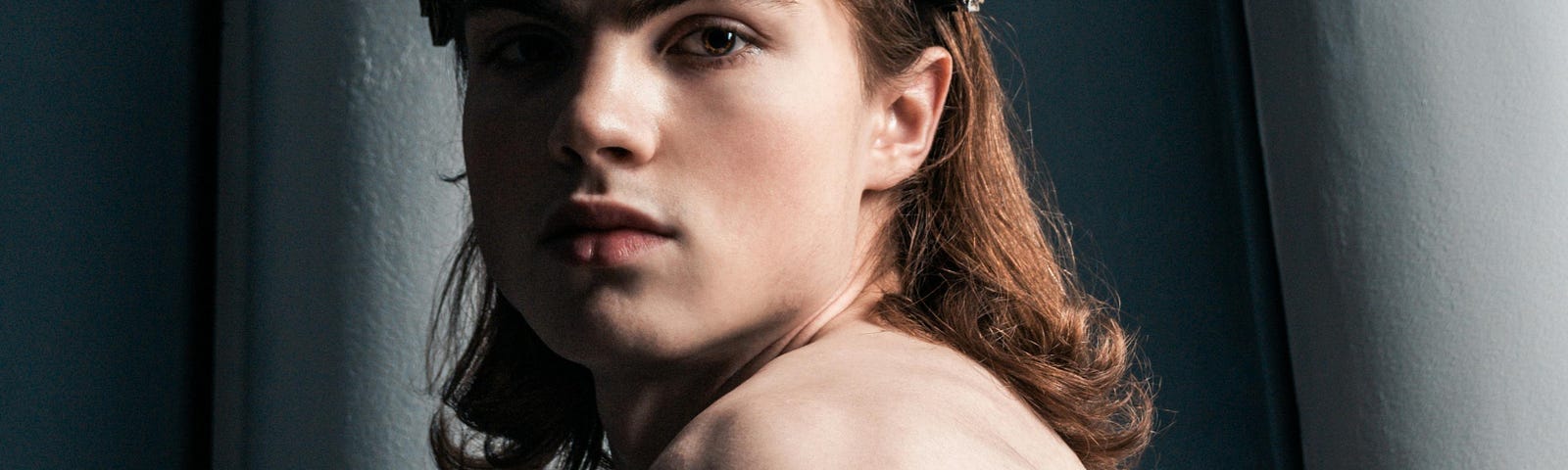 A close-up image of a Caucasian male with long, red hair. He’s wearing a crown of silver skulls, and a single black rose is also visible. He’s shirtless and staring into the camera in an expressionless pose.