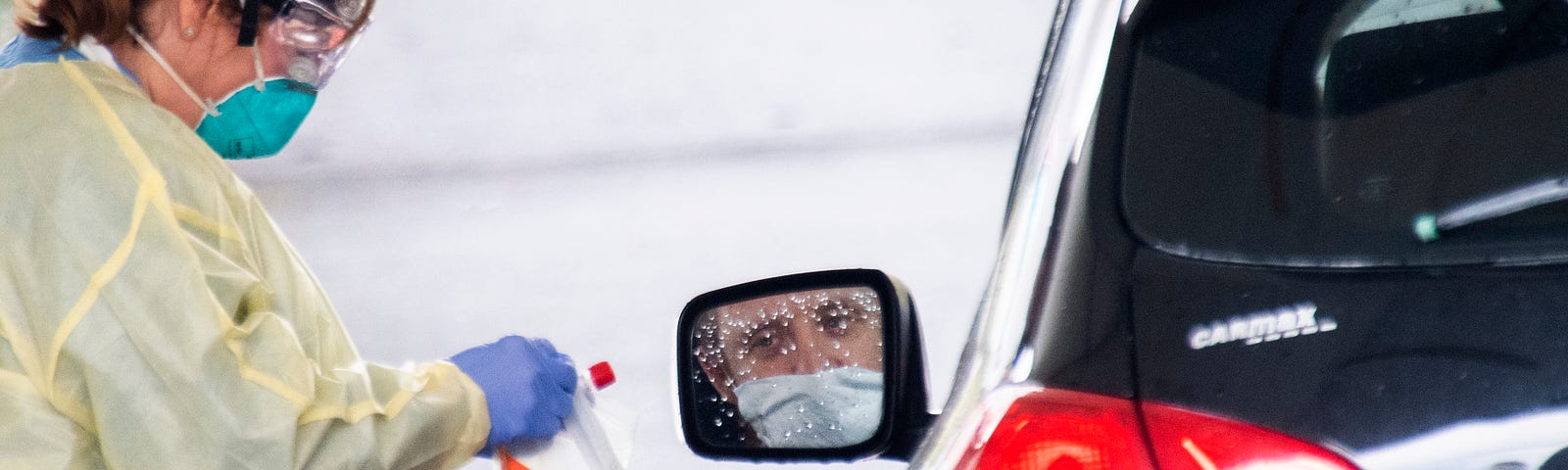 A photo of a man sitting in his car wearing a mask waiting to get tested by a health care worker.