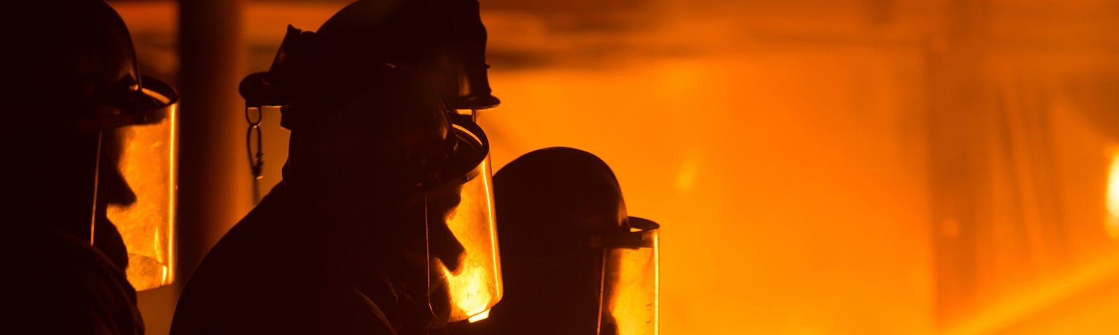 A photo of three firefighters’ shadows illuminated by the orange-red of fires in the background. One of them has a hose.