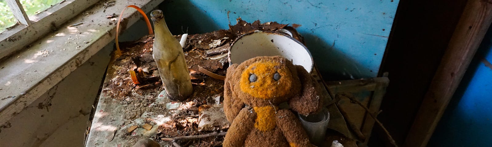 War photo from Ukraine showing a filthy house covered with dirt and debris. On a table is an old stuffed animal, some yellowed papers and some dirty dishes.