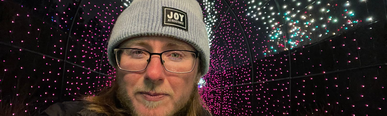 The author is surrounded by white and purple lights. He’s wearing a knitted hat that says “Joy as resistance.” He’s wearing black rimmed glasses and has a beard with enough white to betray that he’s middle-aged. He has long brown hair and a black coat that says “Denver” over a purple sweater. Purple glitter eyeliner matches the lights behind him.