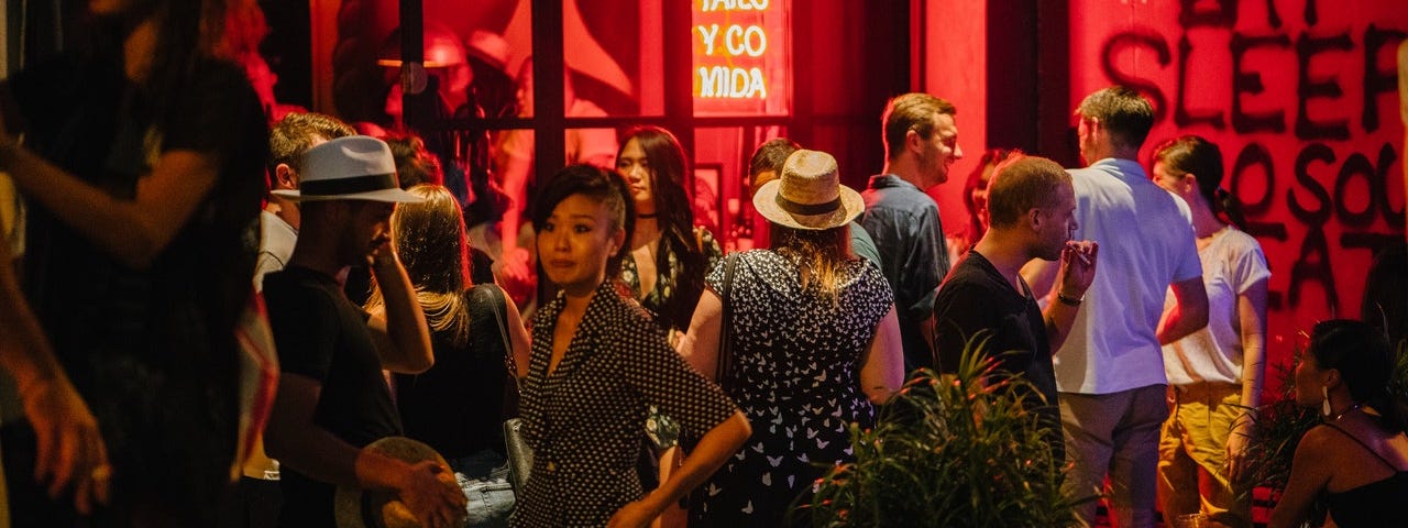 A crowd of people are out the front of a club. One of the girls in the crowd has a comical confused look on her face. The crowd is cast in a nearby neon sign.