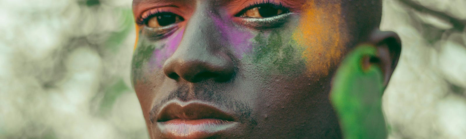 A man of African descent poses outdoors among trees and in-focus leaves. He’s staring at the camera in a close-up, slightly side-on pose to his left. His face is colored with makeup of rainbow hues, and he’s wearing black beads around his bare chest.