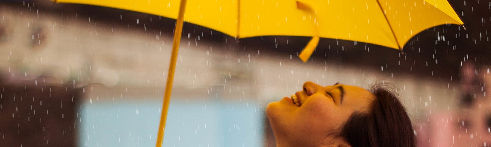 Korean woman stands under a yellow umbrella in the rain with her head tipped back, a big smile on her face