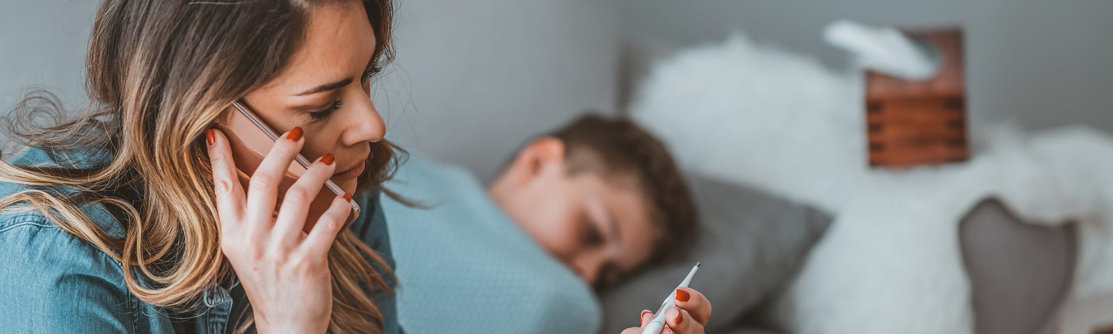 A worried mom on the phone, looking at a thermometer after taking the temperature of her son, who is in bed with a fever.