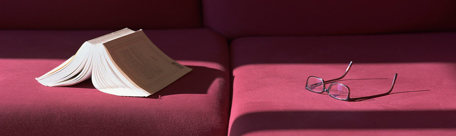 Book and eyeglasses left on a maroon velvet couch.