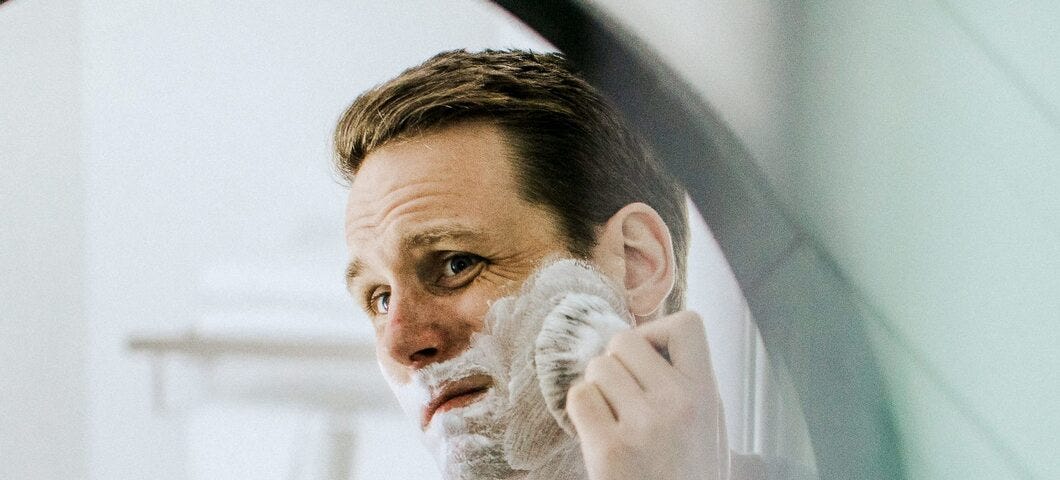 A man looks into a mirror while shaving his face.