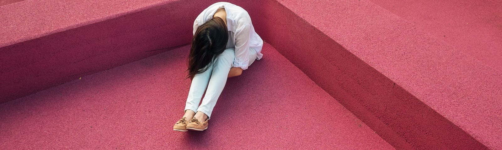 A person with long hair wearing a white shirt and light blue jeans sits with their head to their knees. Dark pink background.