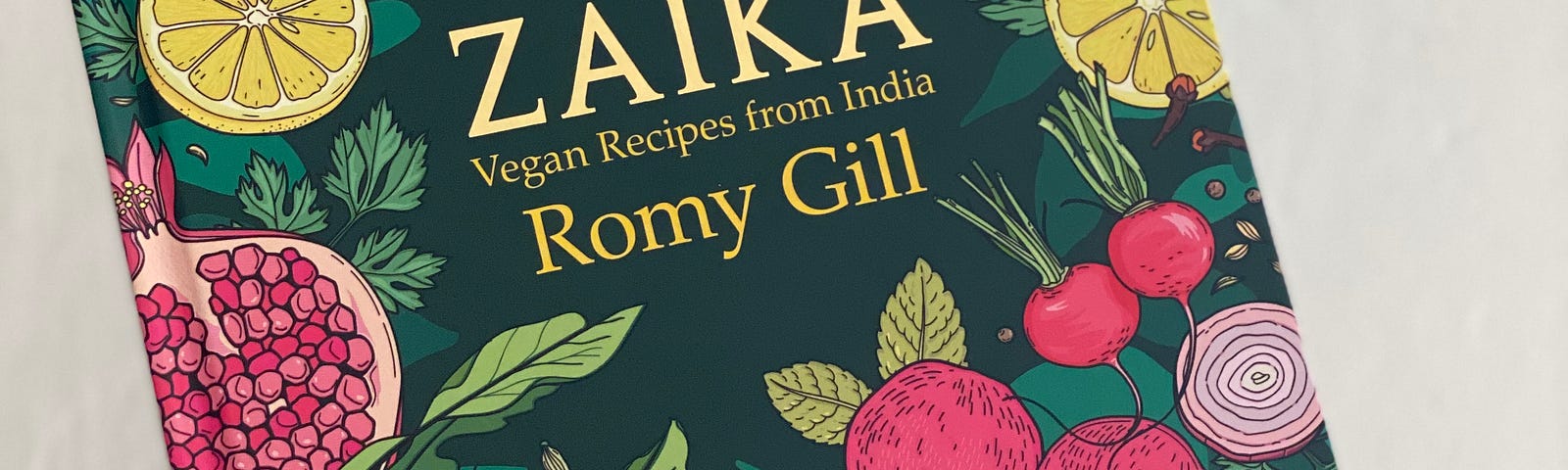 A hand holding the book “Zaika” by Romy Gill, which has illustrations of various vegetables and fruits on a dark green cover