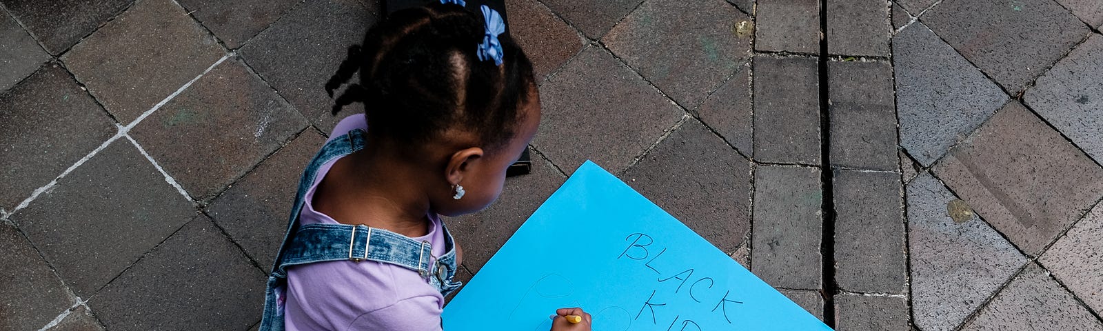 A young Black girl sits on the ground with a bright blue sign that says “Black Kids Matter.”