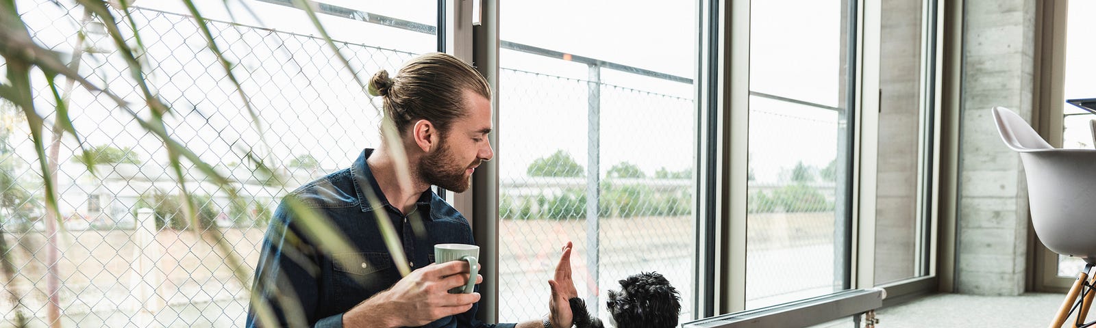 A young man with a laptop sits on the floor in an office and gives a high-five to his dog