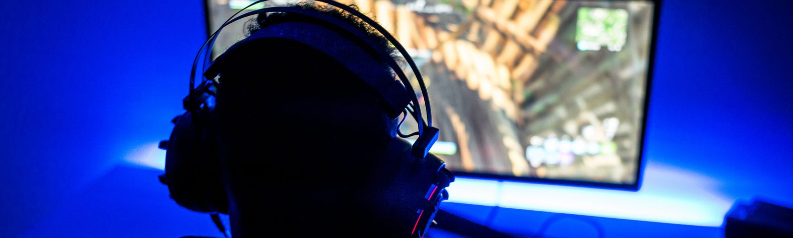 A photo of a teen boy playing video games on a computer. The background is lit with neon blue light and the keyboard is red.