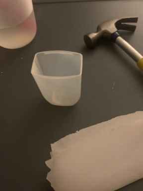 Picture of a clear 2 inch tall bottle cap, a small hammer, a piece of parchment paper, and a water cup.