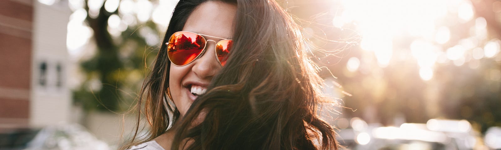 A woman wearing red sunglasses looks to the side and smiles. The sun is glaring behind her.