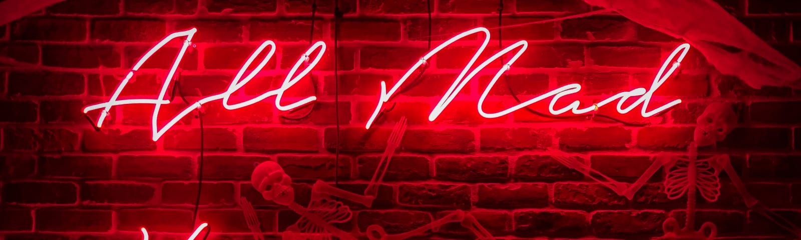 A red neon sign hanging on a brown brick wall. It says “All Mad Here”. A skeleton which looks like a halloween decoration hangs in between in the words.
