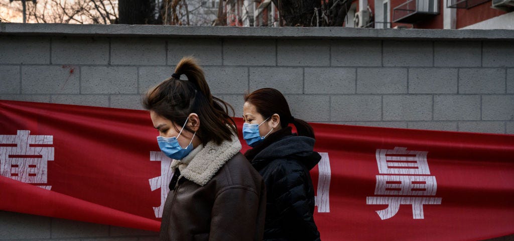 A photo of two Chinese women wearing protective masks as they pass a red propaganda banner.