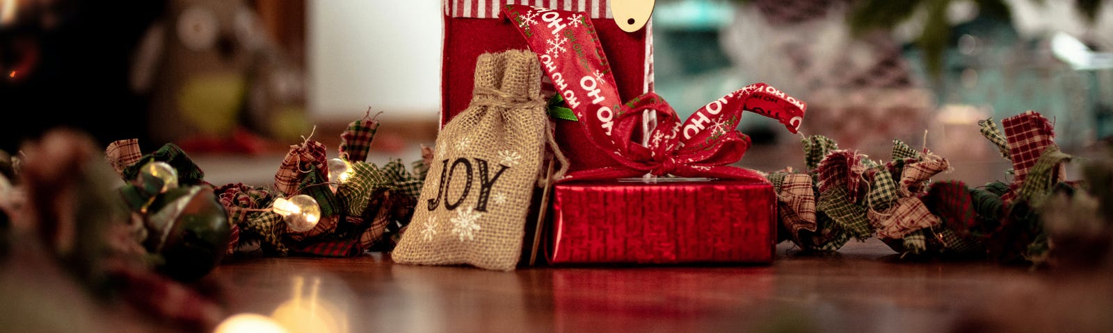 A small burlap bag with the word joy written on it leans against a red wrapped gift and a red gift bag with bright polka-dotted paper. A Christmas tree is in the background slightly out of focus.
