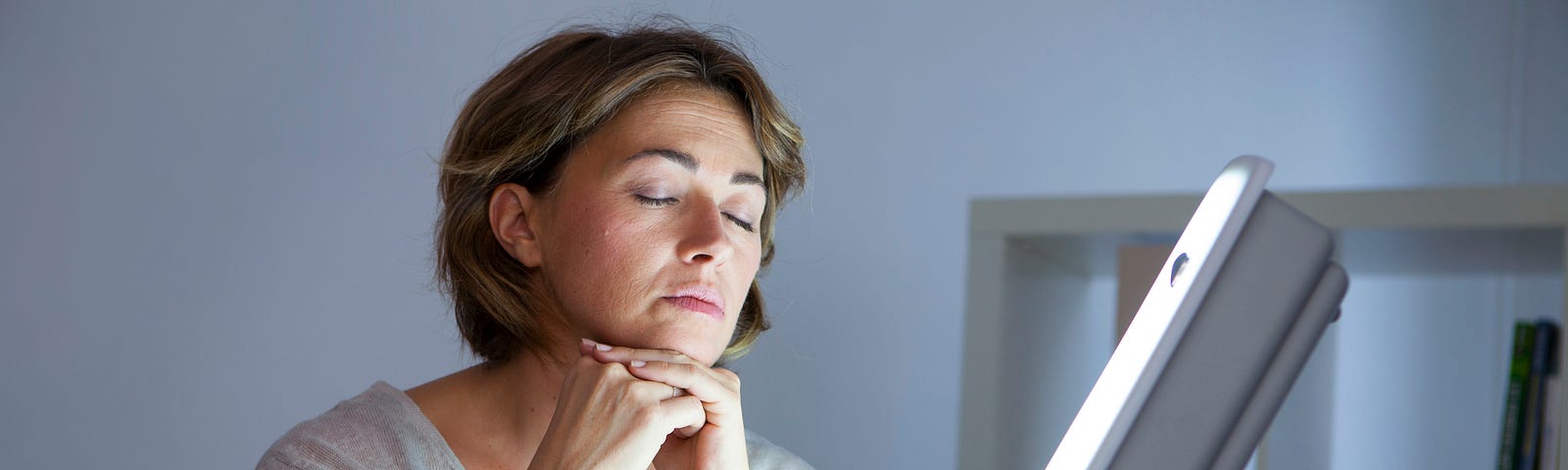 A tired-looking person sitting in front of a large rectangular SAD lamp, resting their chin on their hands.