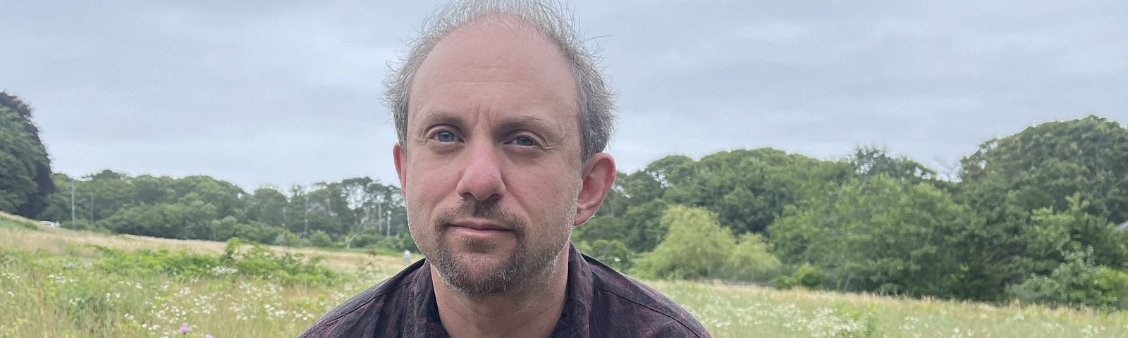 Tucker Lieberman, age 44, bald and with a goatee, crouched in a grassy meadow with white flowers and trees in the background. Wearing a collared shirt, red exercise pants, sneakers.