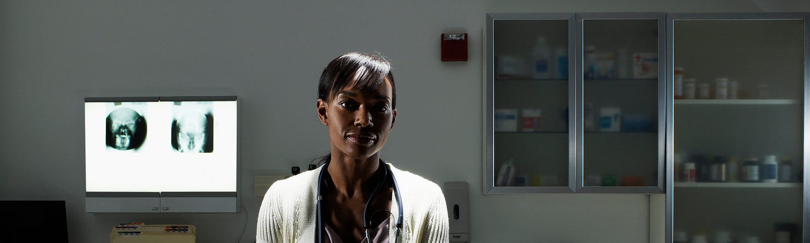 A photo of a Black female doctor sitting in an examination room, arms crossed.