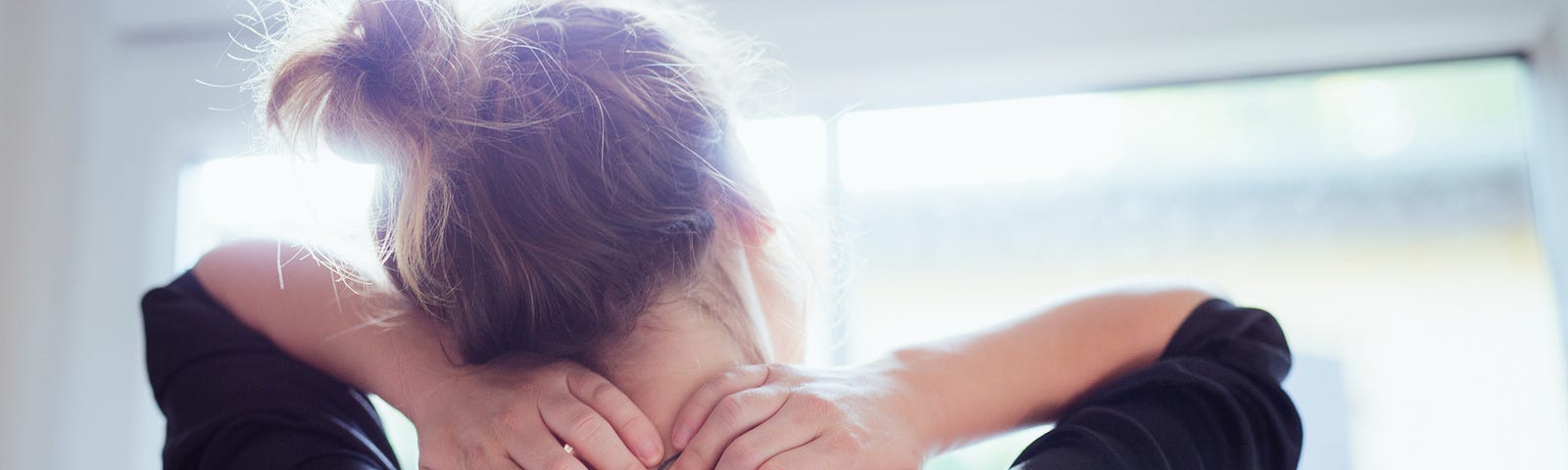 Rear view of a young woman looking out the window as she massages her neck.