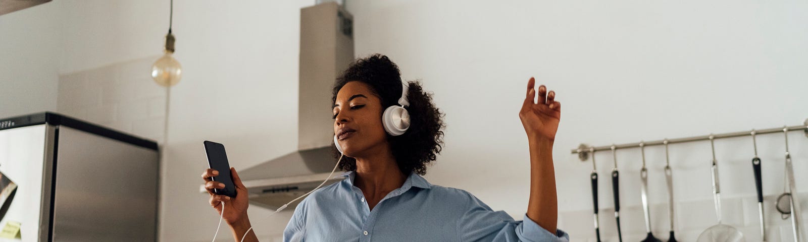 A photo of a black woman listening to music on headphones in her kitchen. She is feeling herself.