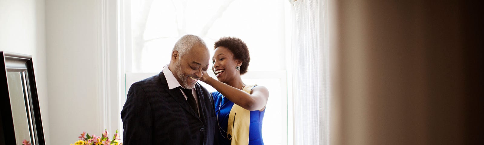 A Black woman assists her elder father with his collar as they prepare for a party.