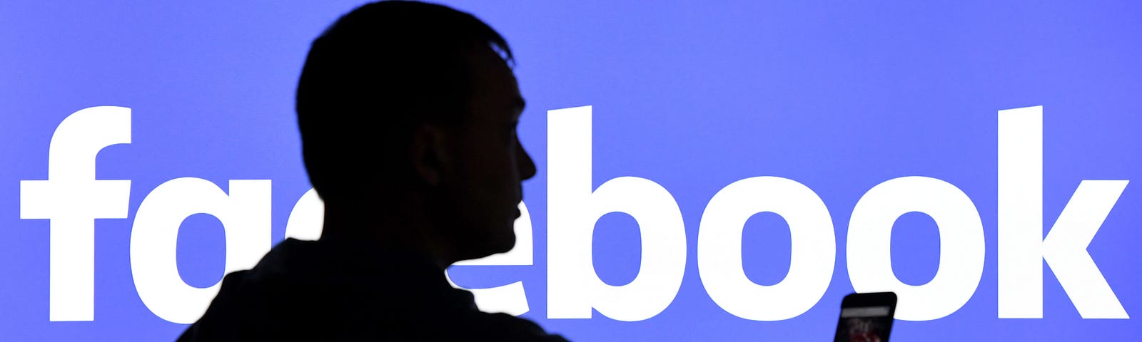 A man with a smartphone stands in front of a monitor with the Facebook logo.