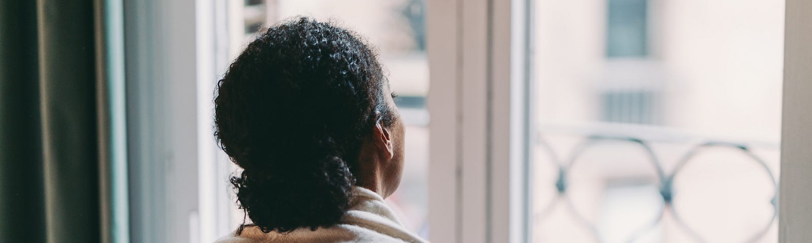 Black woman holding a cup of coffee while looking out her window.