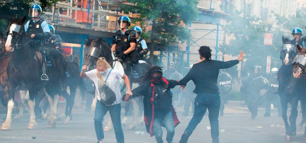 A photo of police tear gassing protestors as the police ride on horses past them.