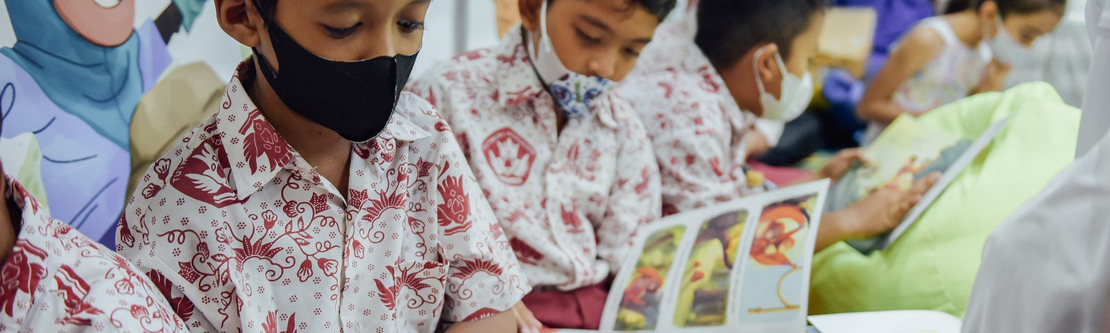 Three boys wearing matching white shirts with a red pattern and masks reading children’s books in a classroom setting.