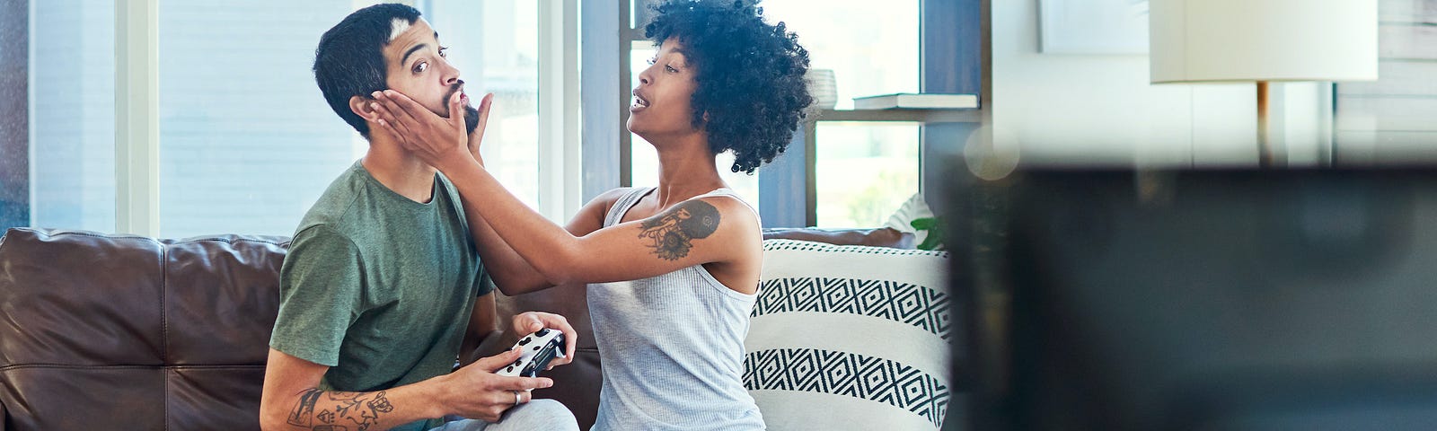 A couple sitting on a couch. One has their hands on the face of the other, who is distracted playing video games.