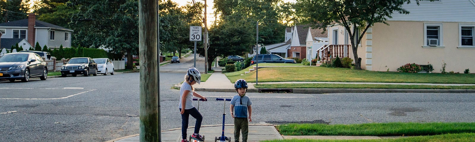 Two children stand on a residential street corner with their scooters.