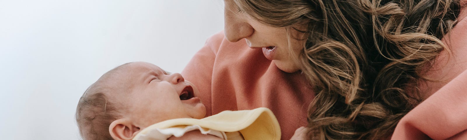 Woman wearing salmon-coloured sweater looking down at crying infant in her arms.