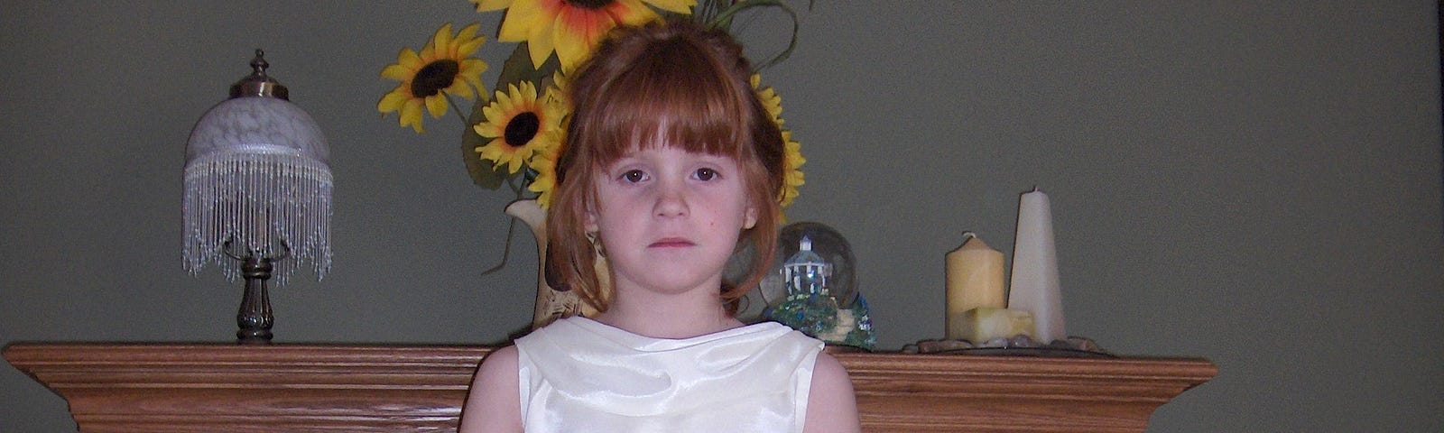 Young Fiona stands in front of a fireplace mantle in a white sleeveless dress. She is not smiling.